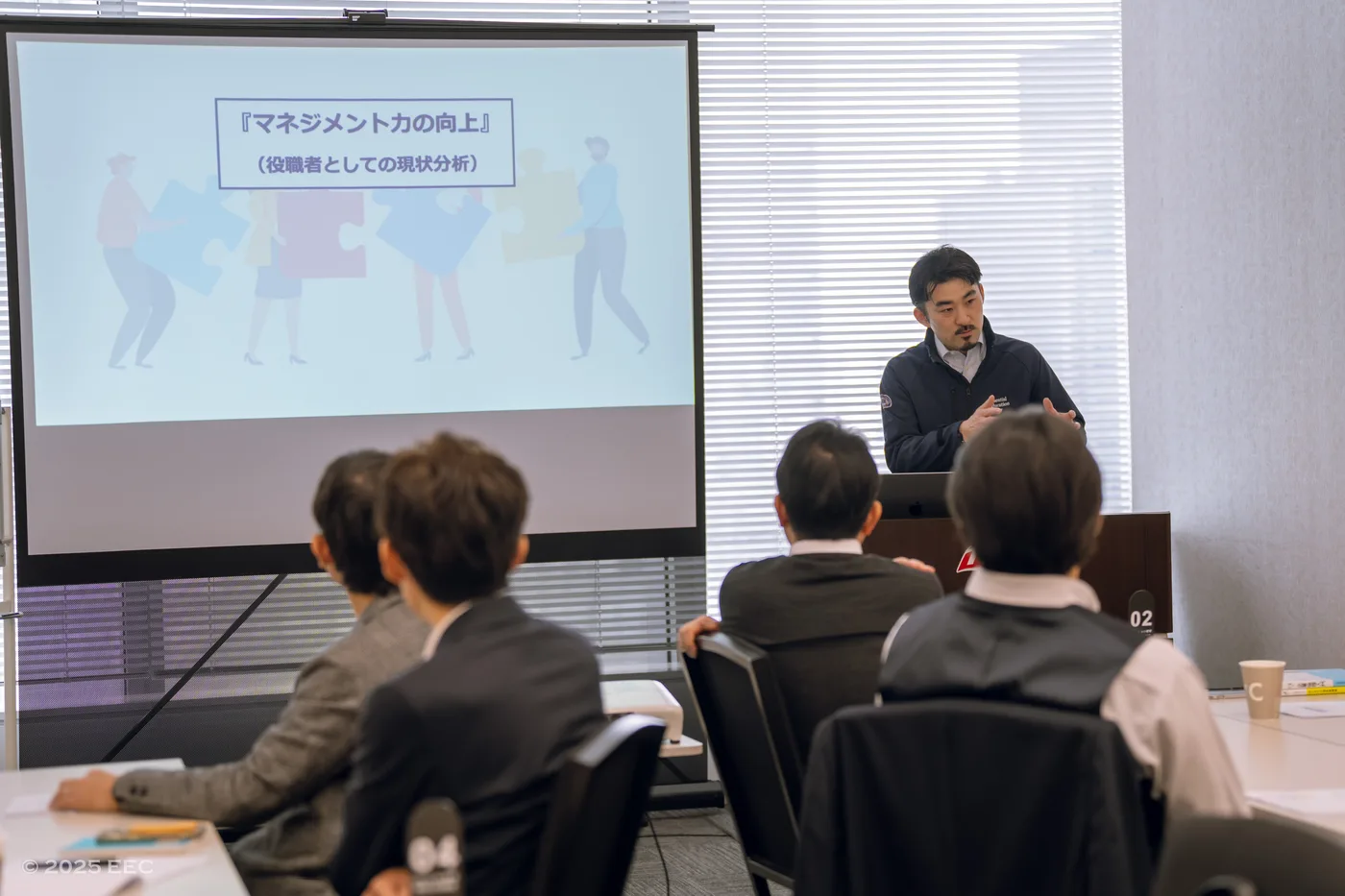 エッセンシャルエデュケーションセンターの代表である田中がプレゼン中。スライドには「マネジメント力の向上」の文字。参加者数名が座って聞いている。明るい雰囲気。